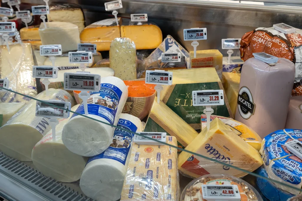 Assorted gourmet cheeses in a refrigerated deli display case featuring electronic shelf labels and digital price tags for real-time pricing.
